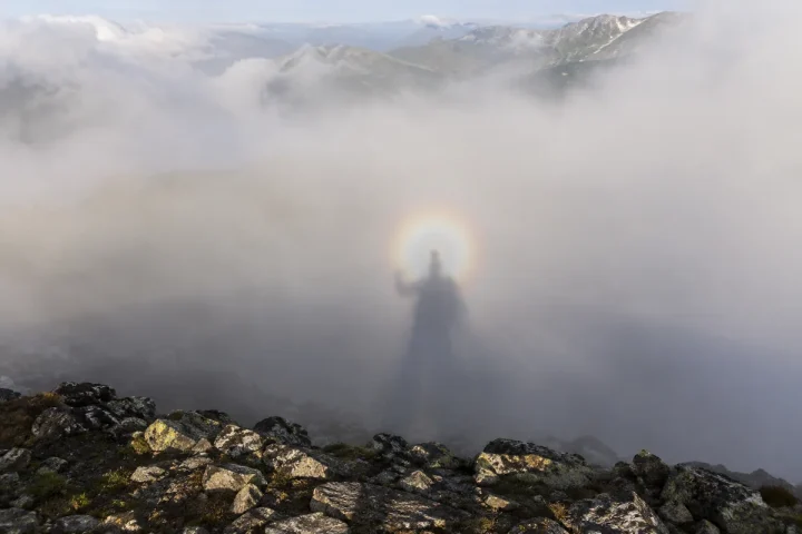Cień obserwatora z uniesioną ręką na górskiej mgle, z subtelnymi pierścieniami wokół cienia głowy